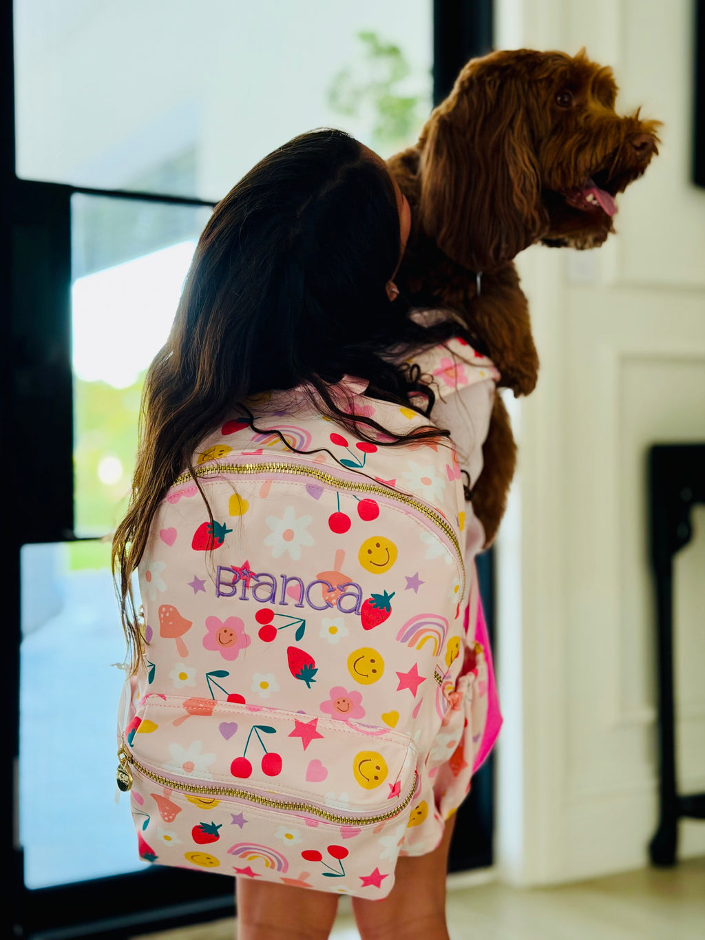 Girl wearing a pink backpack with cherries, smiley faces, and rainbows, custom embroidered with the name Bianca, while hugging her dog
