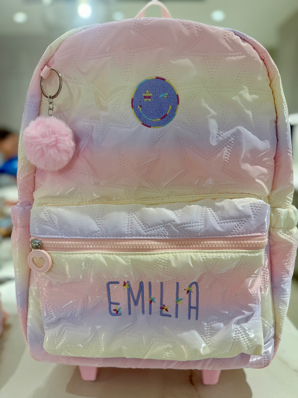 Quilted pastel rainbow rolling backpack with a pink pom pom, custom embroidered with the name Emilia in purple letters with tiny flower accents