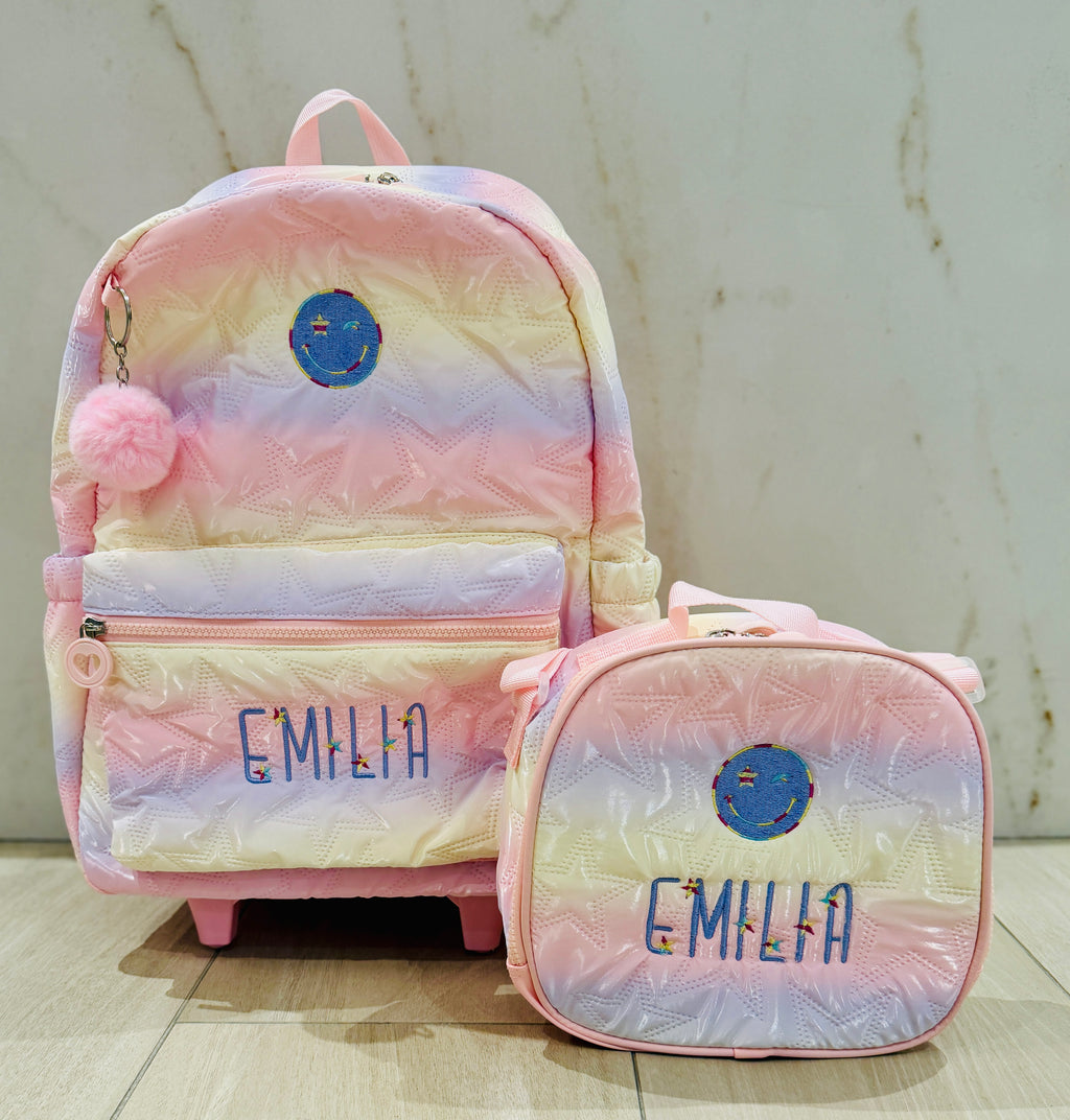 Pastel rainbow rolling backpack and matching lunch box set, each custom embroidered with the name Emilia in purple letters with tiny flower accents