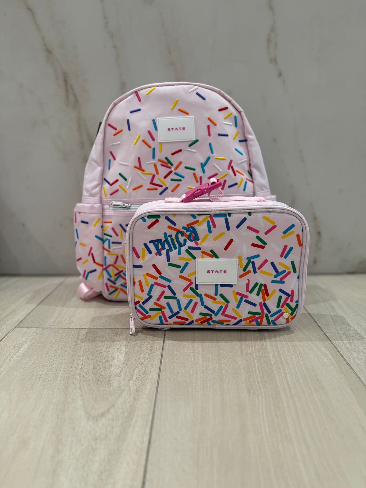 Pink rainbow sprinkles backpack and lunch box set with the name Mica custom embroidered in blue script